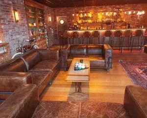 Gezellige bar met lederen zithoeken, houten tafels en verlichte drankkast tegen bakstenen muur in warme sfeer